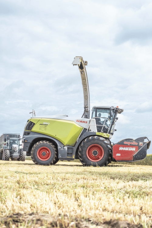 Grande machine agricole verte stationnée dans un champ moissonné sous un ciel nuageux.