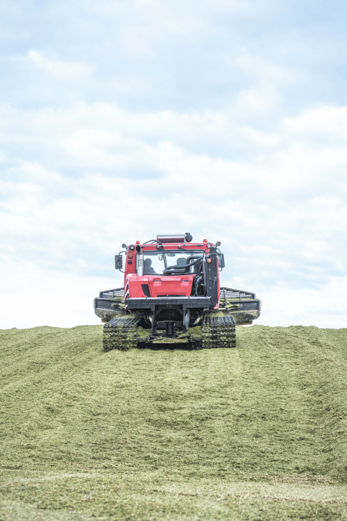 Véhicule agricole rouge à chenilles sur champ de foin, ciel nuageux en arrière-plan.