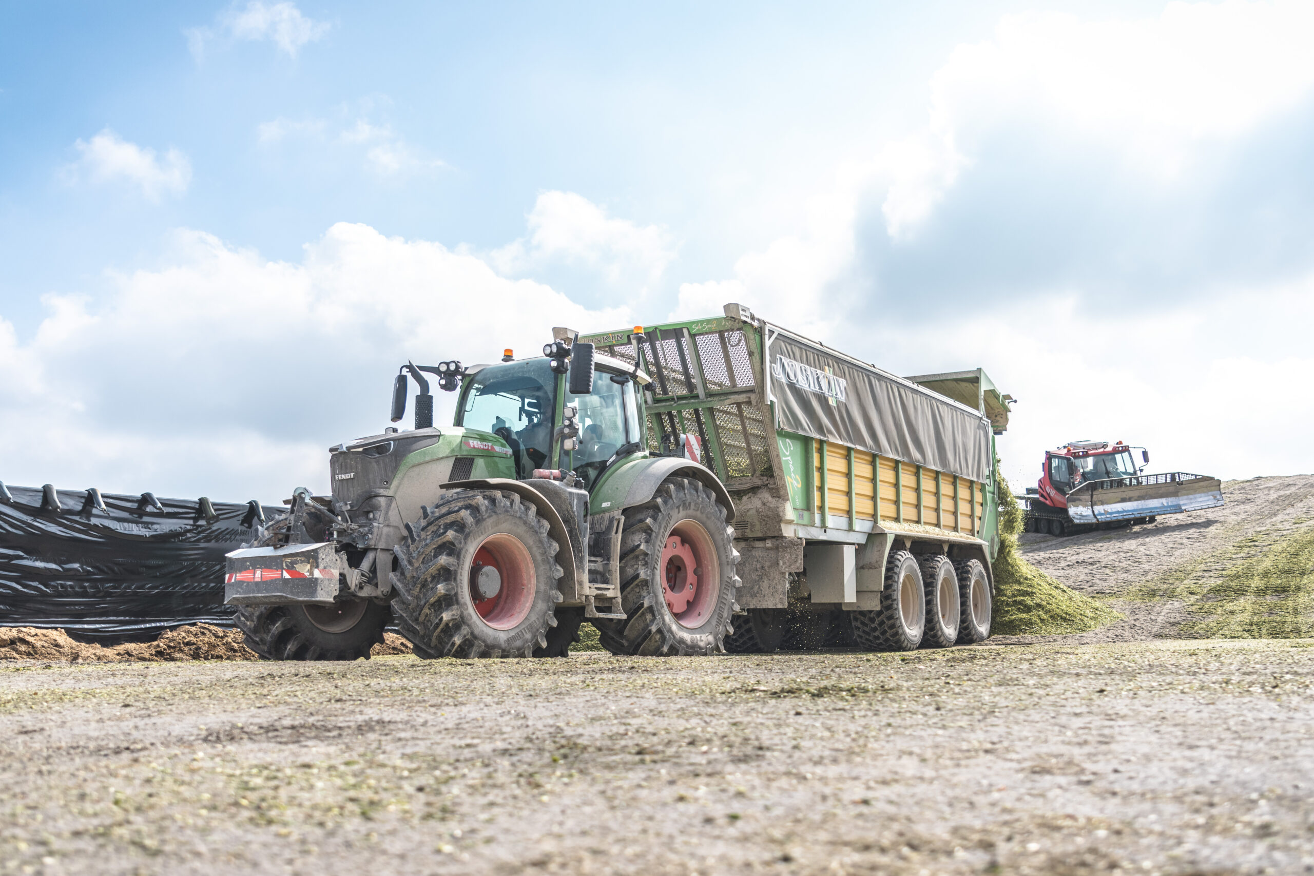 Véhicule agricole déchargeant du fourrage haché devant des silos industriels en plein air.
